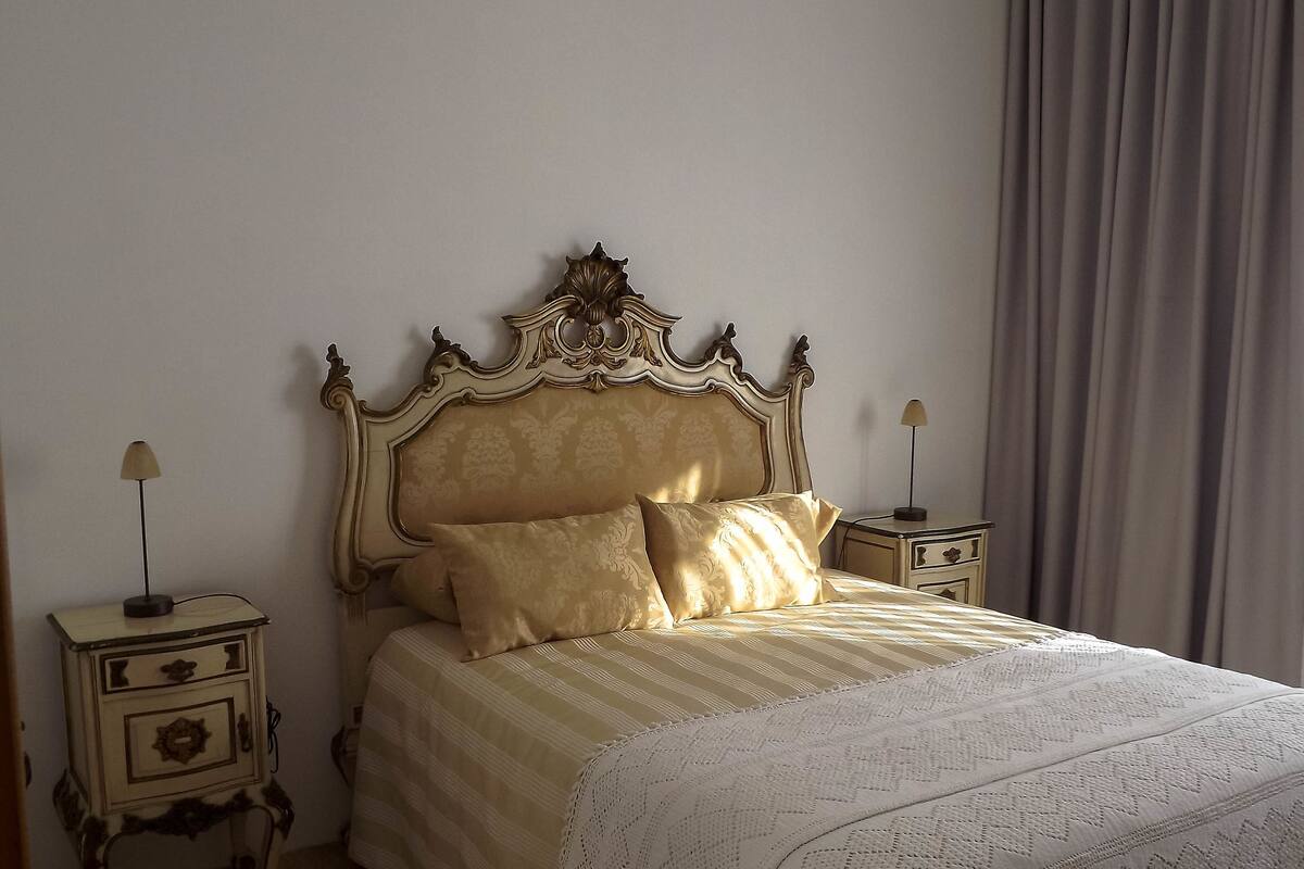 A bedroom with a classic bed, two bedside tables, a lamp on each, and a large window with curtains.