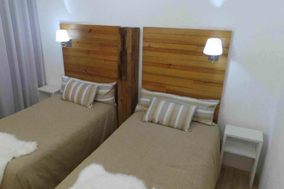 A room with two single beds, each with a wooden headboard and bedside lamps. There is a white nightstand on each side of the beds.