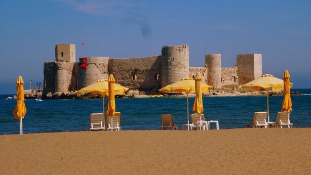 A beach with yellow umbrellas and white lounge chairs, a stone castle in the background, and clear blue water. - Eylul Hotel (Erdemli)