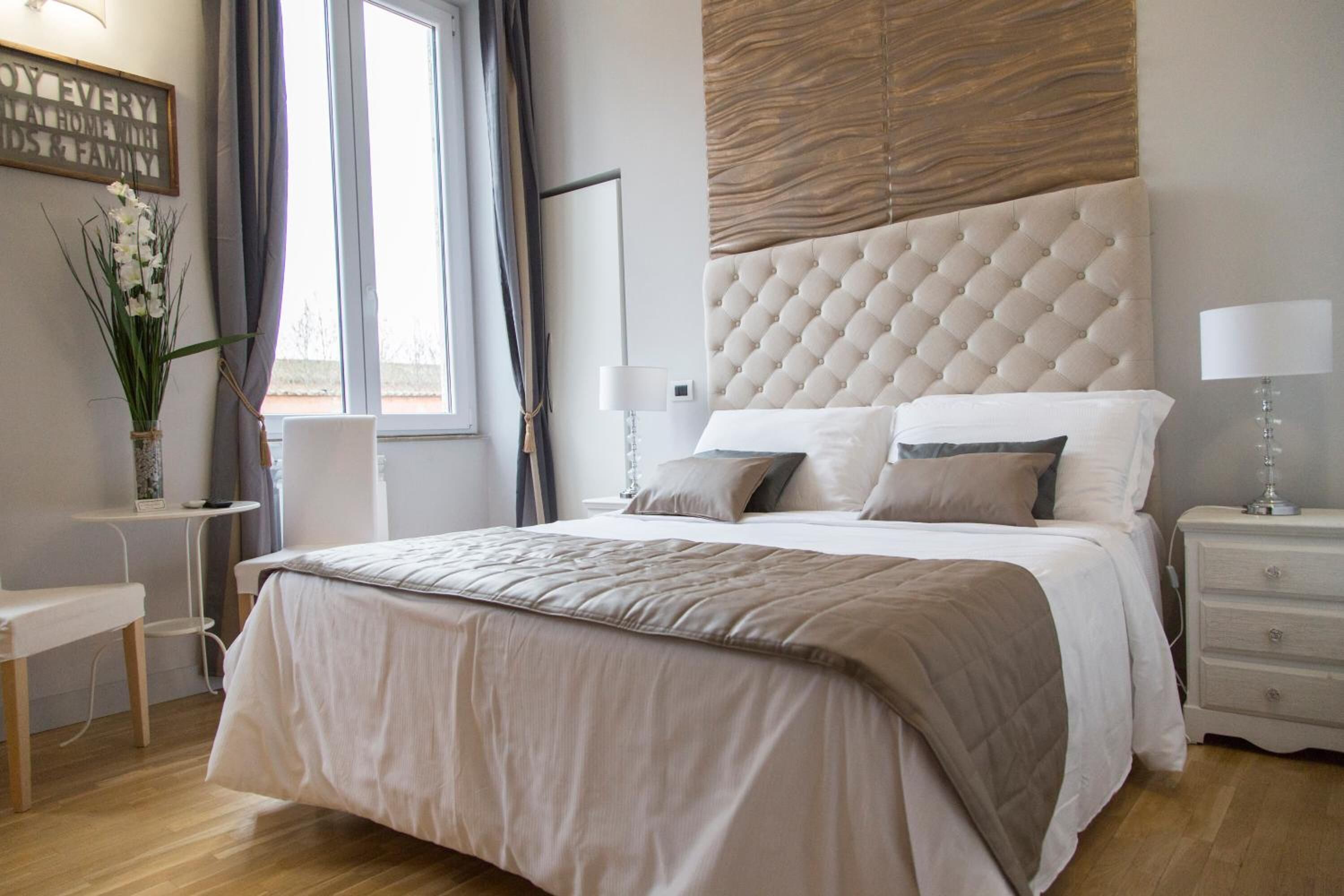 A modern bedroom with a large bed, a bedside table, a nightstand, a wall-mounted lamp, and a window with curtains.