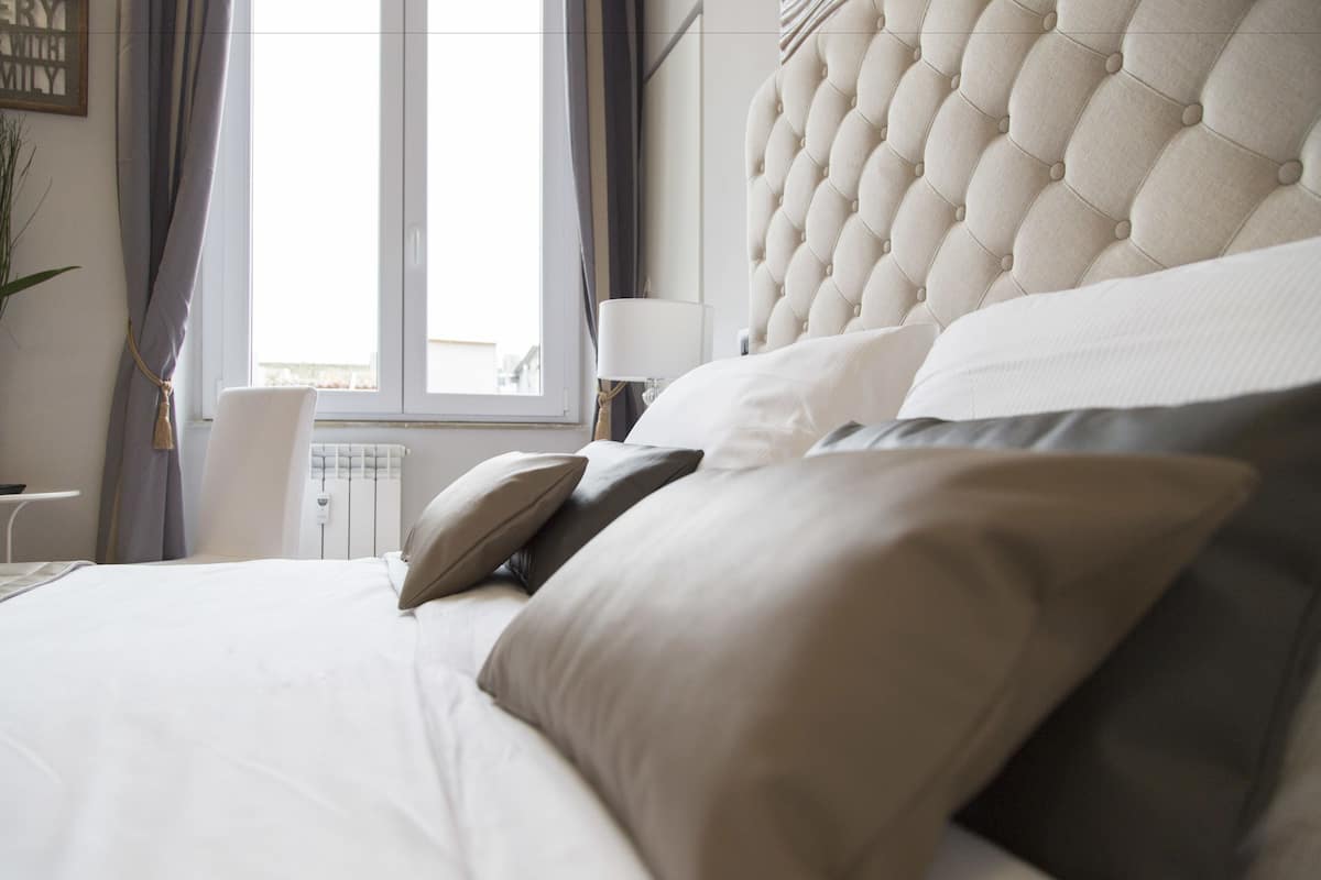Una cama bien hecha con un cabecero capitonado, una mesita de noche con una lámpara y vistas al exterior a través de una ventana.