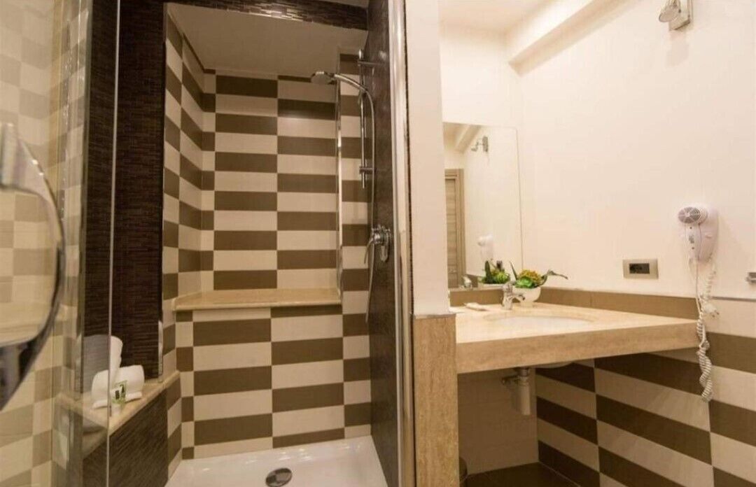 A bathroom with a checkered tile wall, a glass shower enclosure, a sink with a mirror, and a hairdryer on the wall.