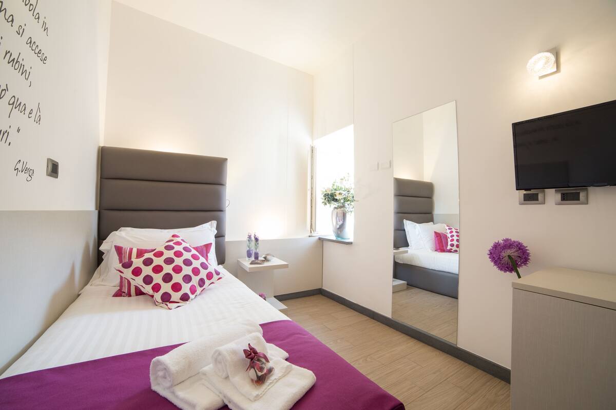 A modern hotel room with a large bed, a bedside table, a mirror, a TV, and a small cabinet.