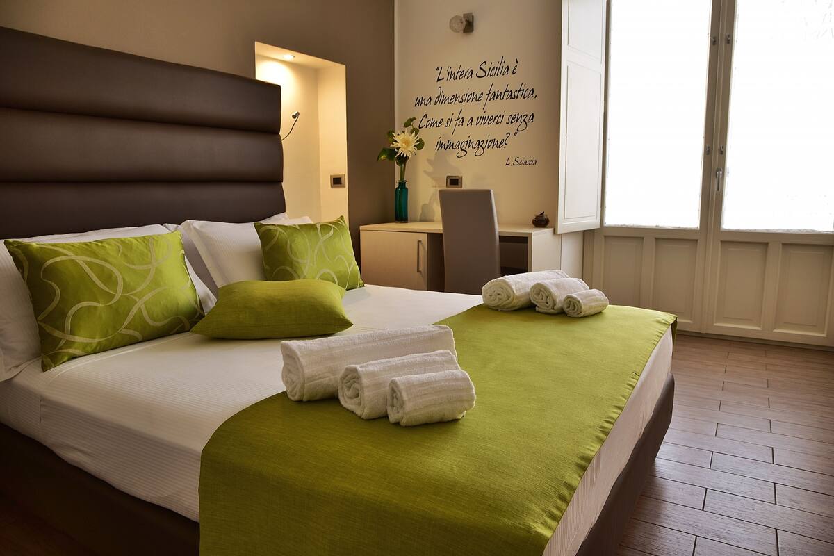 A neatly made bed with green and white pillows, a green bedspread, and rolled white towels.