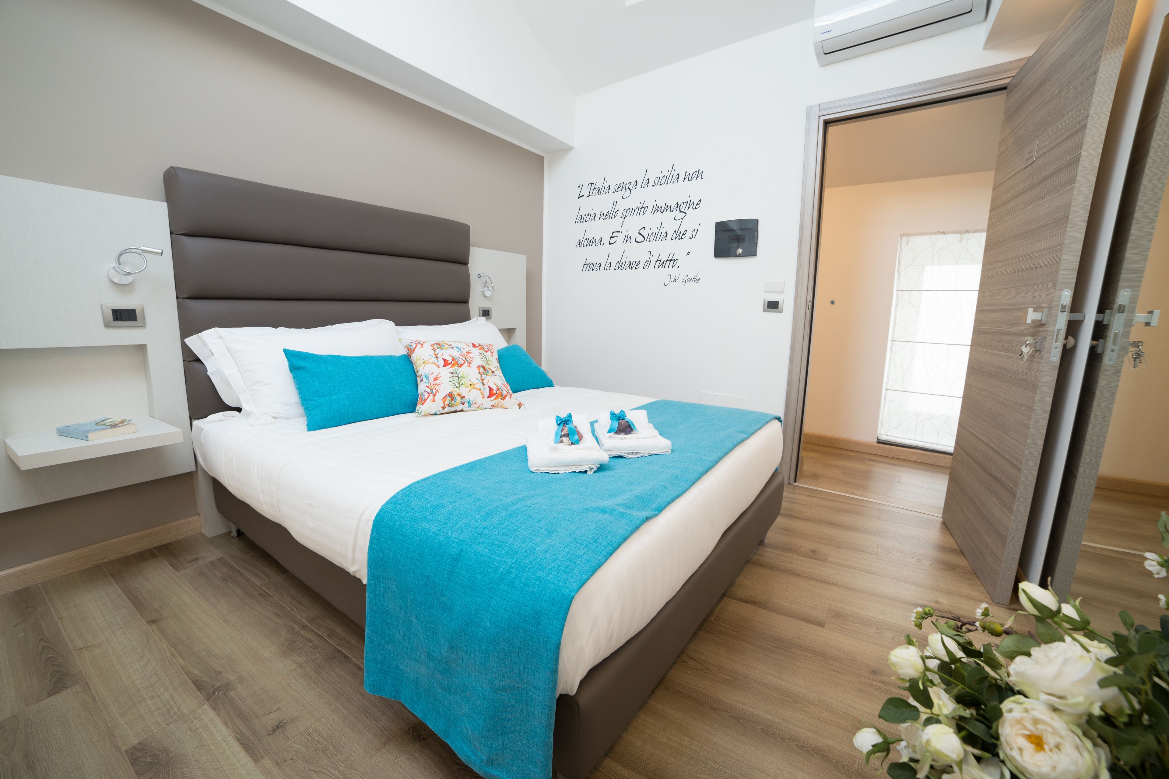 Un dormitorio moderno con una cama grande, una mesita de noche, una lámpara de pared y una puerta que da a otra habitación.