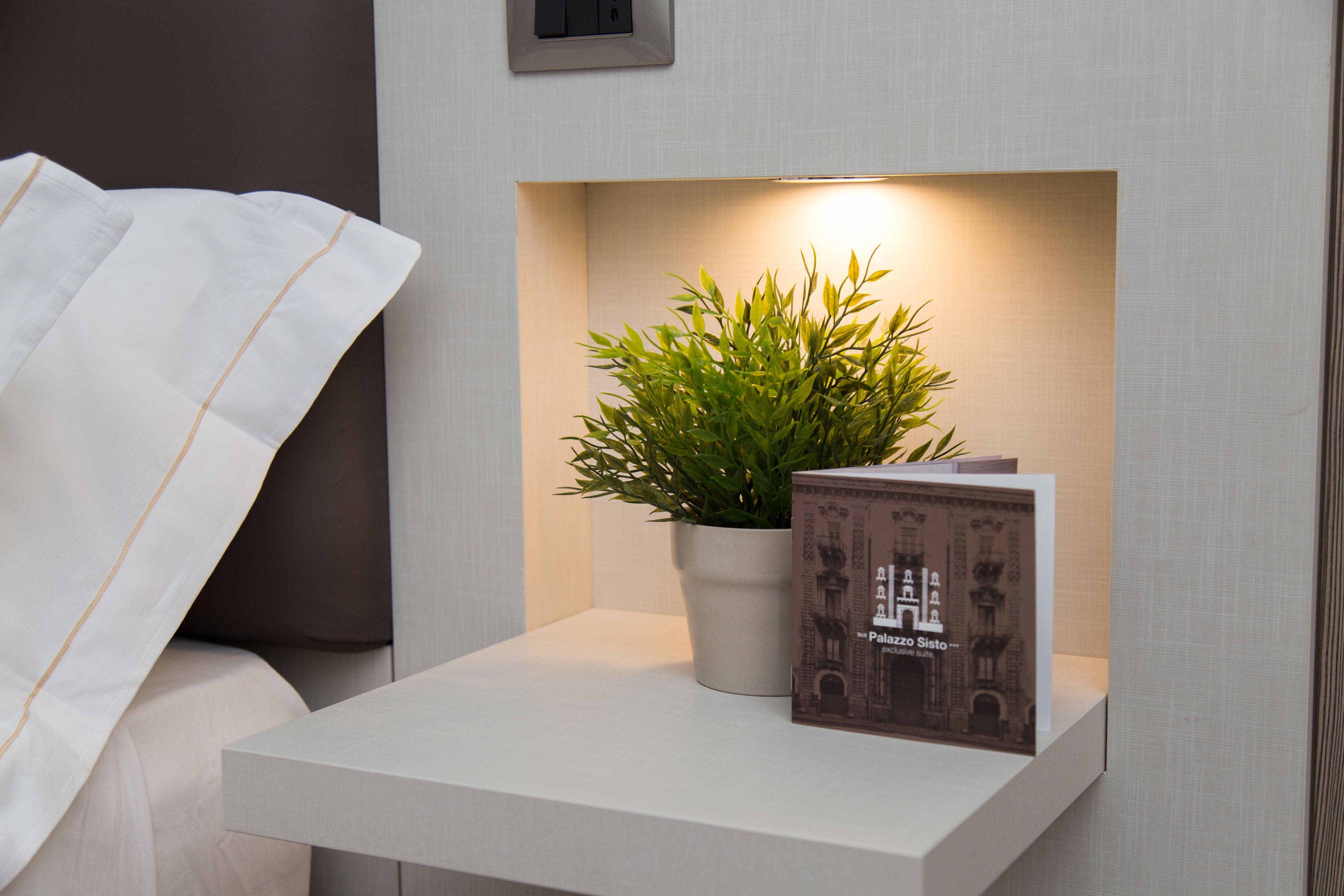 A white bedside table with a potted plant and a book titled "Palazzo Sisto."