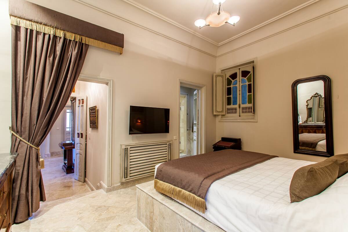 Un dormitorio con cama, televisor, espejo y puerta que da a otra habitación.