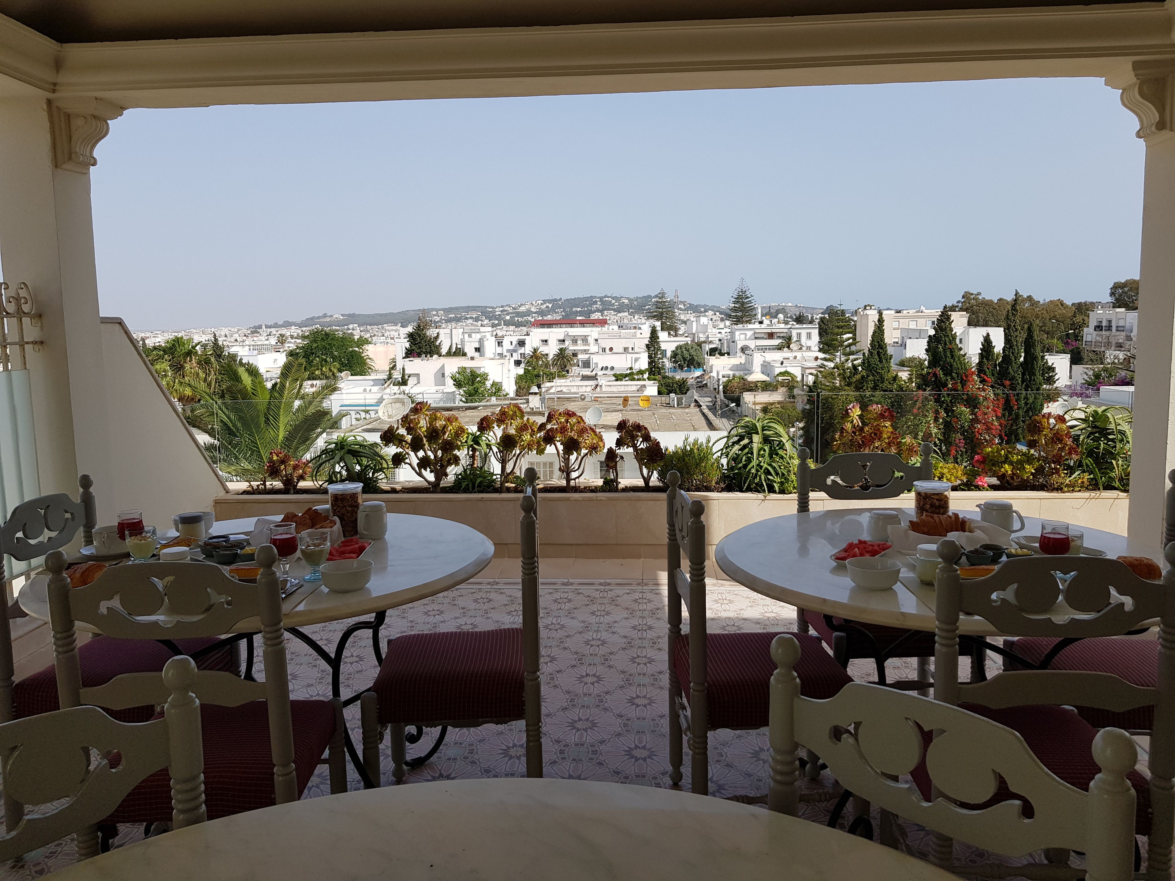 A terrace with two round tables and chairs, overlooking a cityscape with buildings and greenery.
