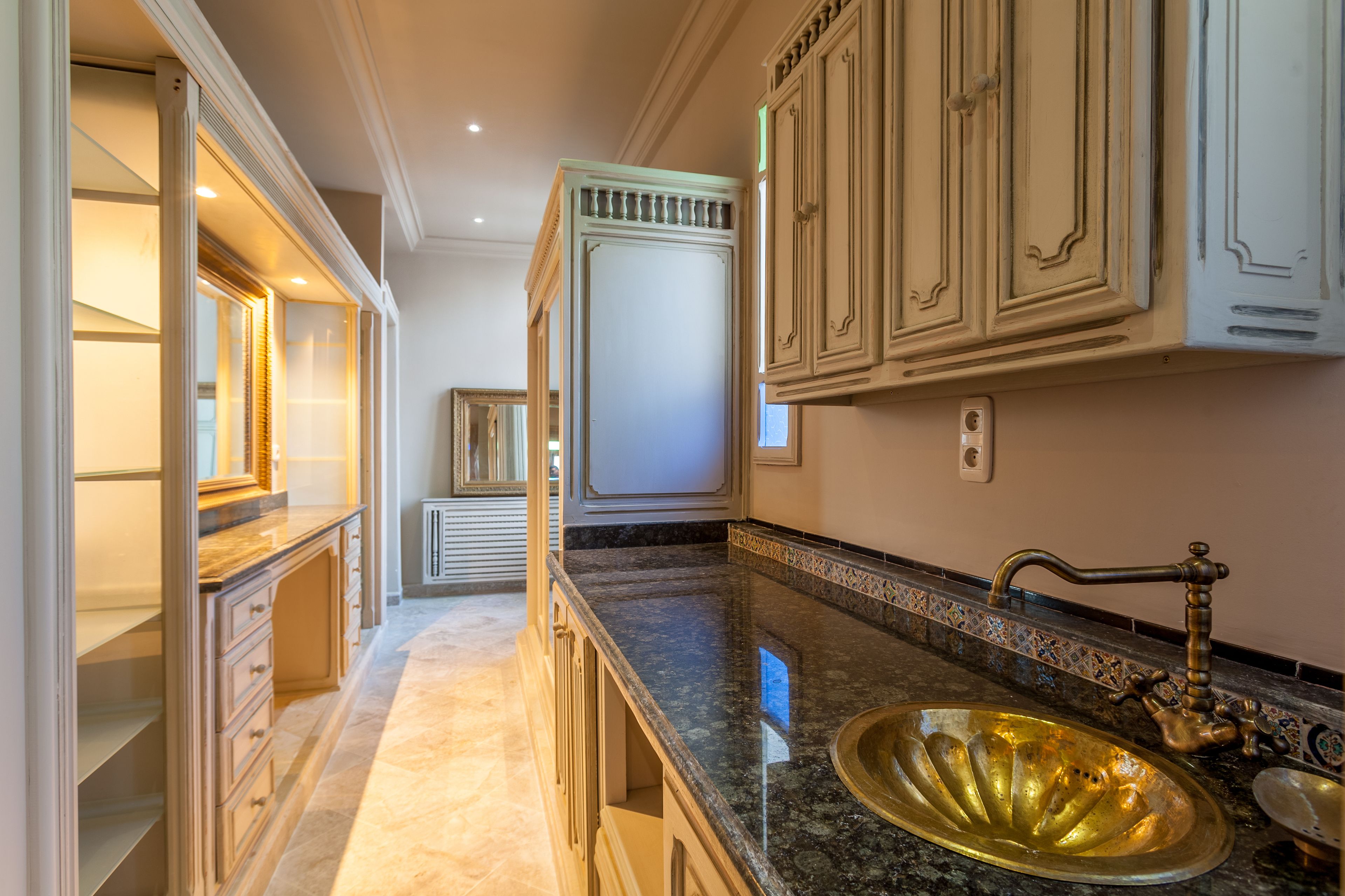 Una cocina con encimera de mármol, pileta dorada y muebles blancos.