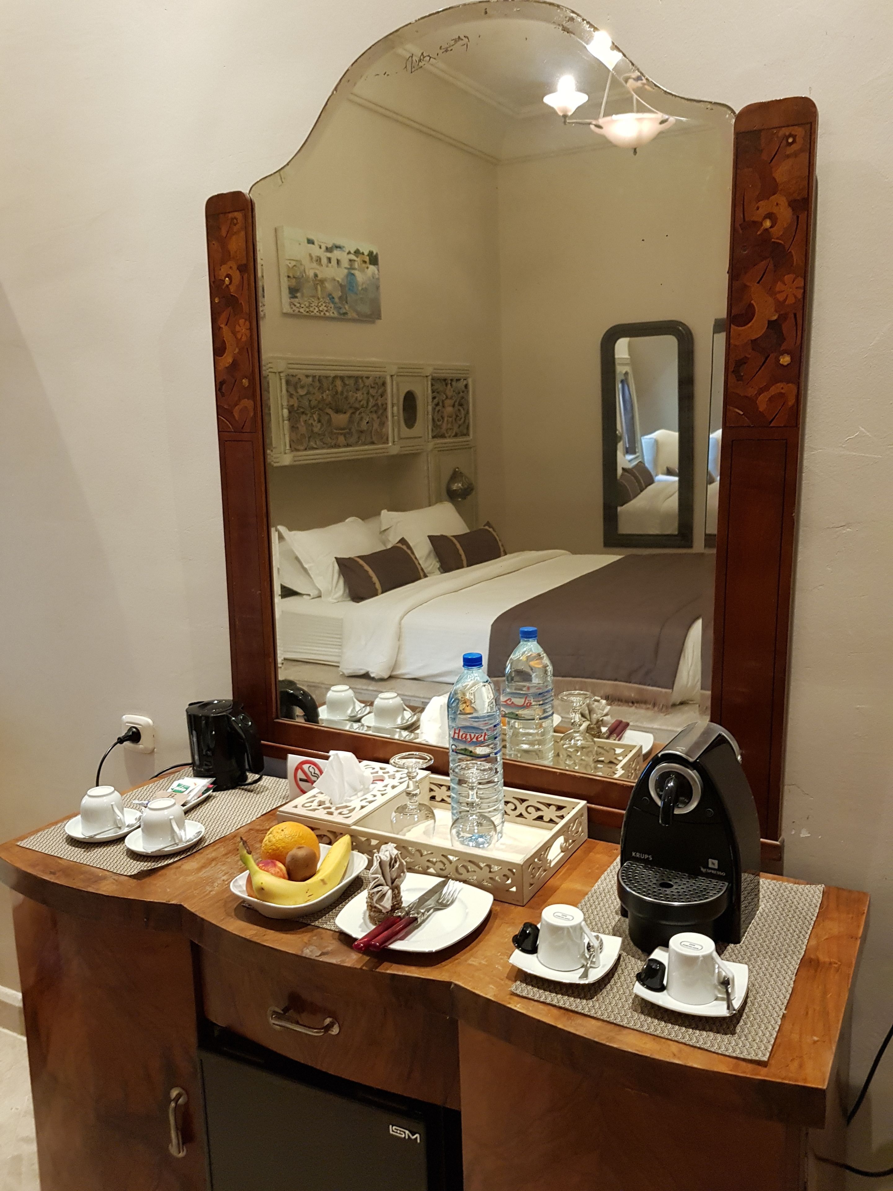 A hotel room with a wooden dresser, a mirror, a bed with white bedding, a coffee maker, and a tray with breakfast items.