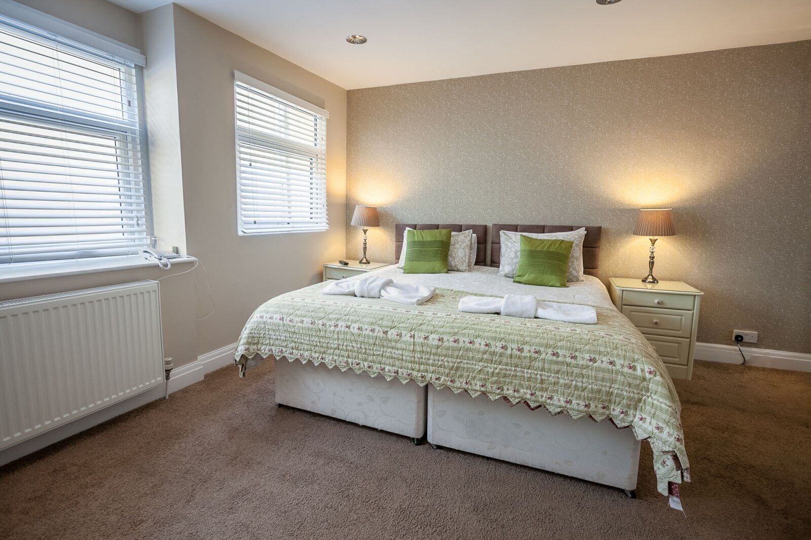 A bedroom with a bed, two bedside lamps, a radiator, and two windows with blinds.