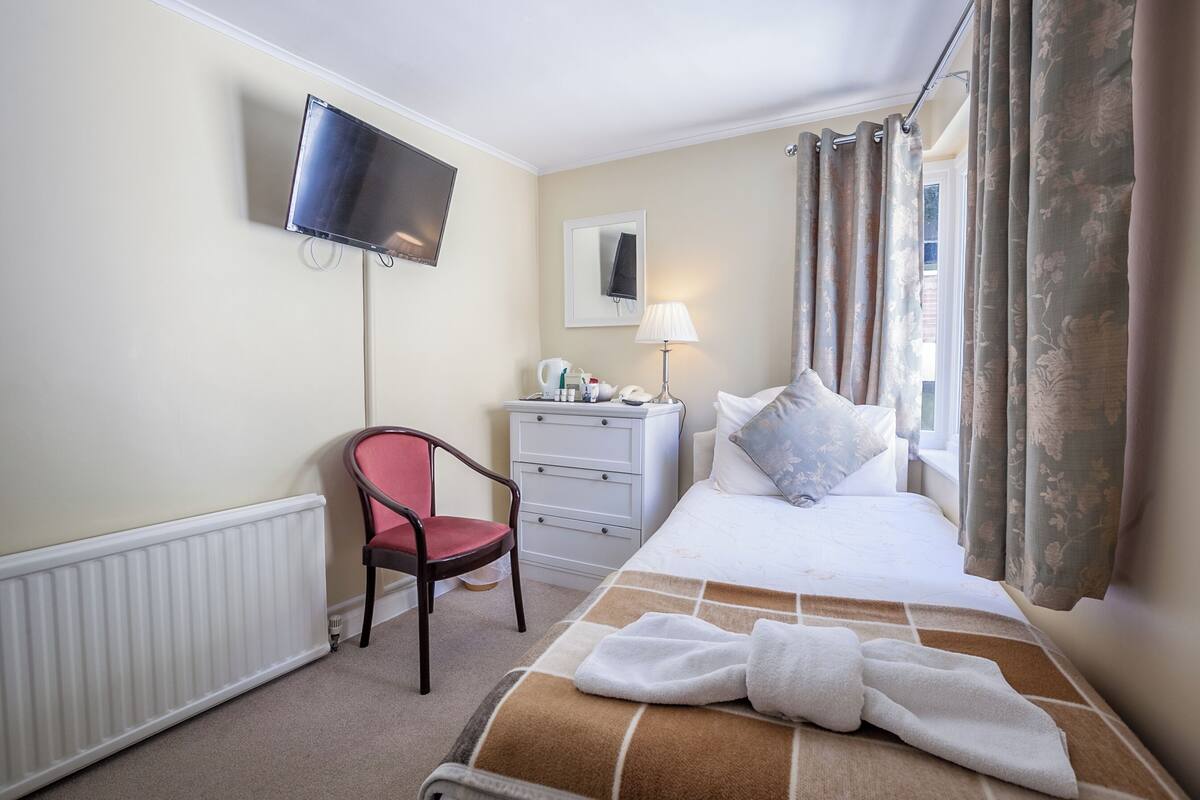 Quarto de hotel com cama, poltrona, cômoda, televisão e janela com cortinas.