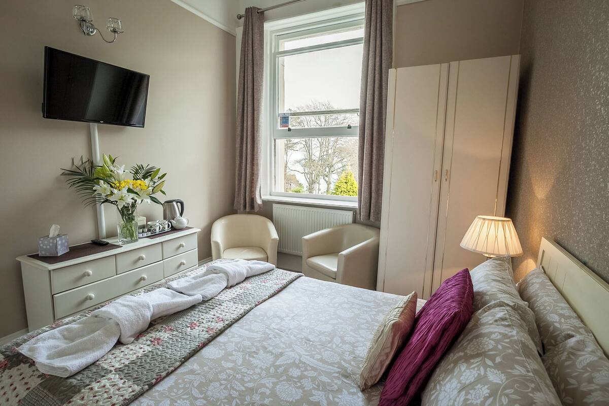 Quarto de hotel com cama, televisão na parede, escrivaninha com um vaso de flores, cadeira e guarda-roupa.