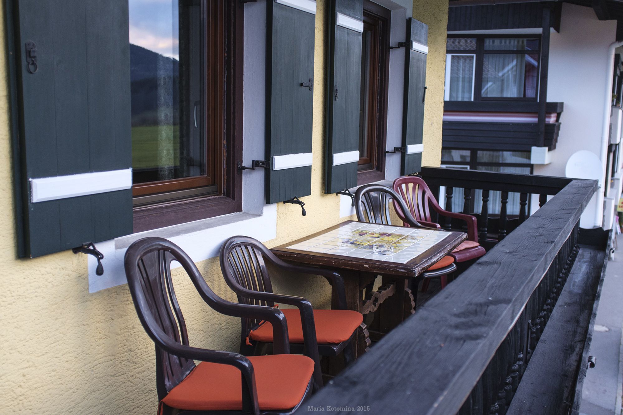 A wooden table with chairs on a balcony, overlooking a building with large windows and a railing.
