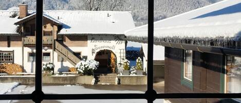 Vista a través de una ventana con un diseño de cuadrícula, mostrando un edificio con un letrero que dice "Landhaus Sunnmatt" y un paisaje montañoso nevado al fondo.