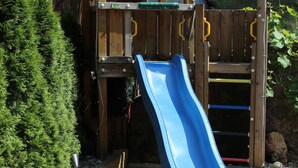 A wooden play structure with a blue slide and a climbing area, surrounded by a wooden fence and greenery. - Hotel Pension Almrose (Wildschönau)