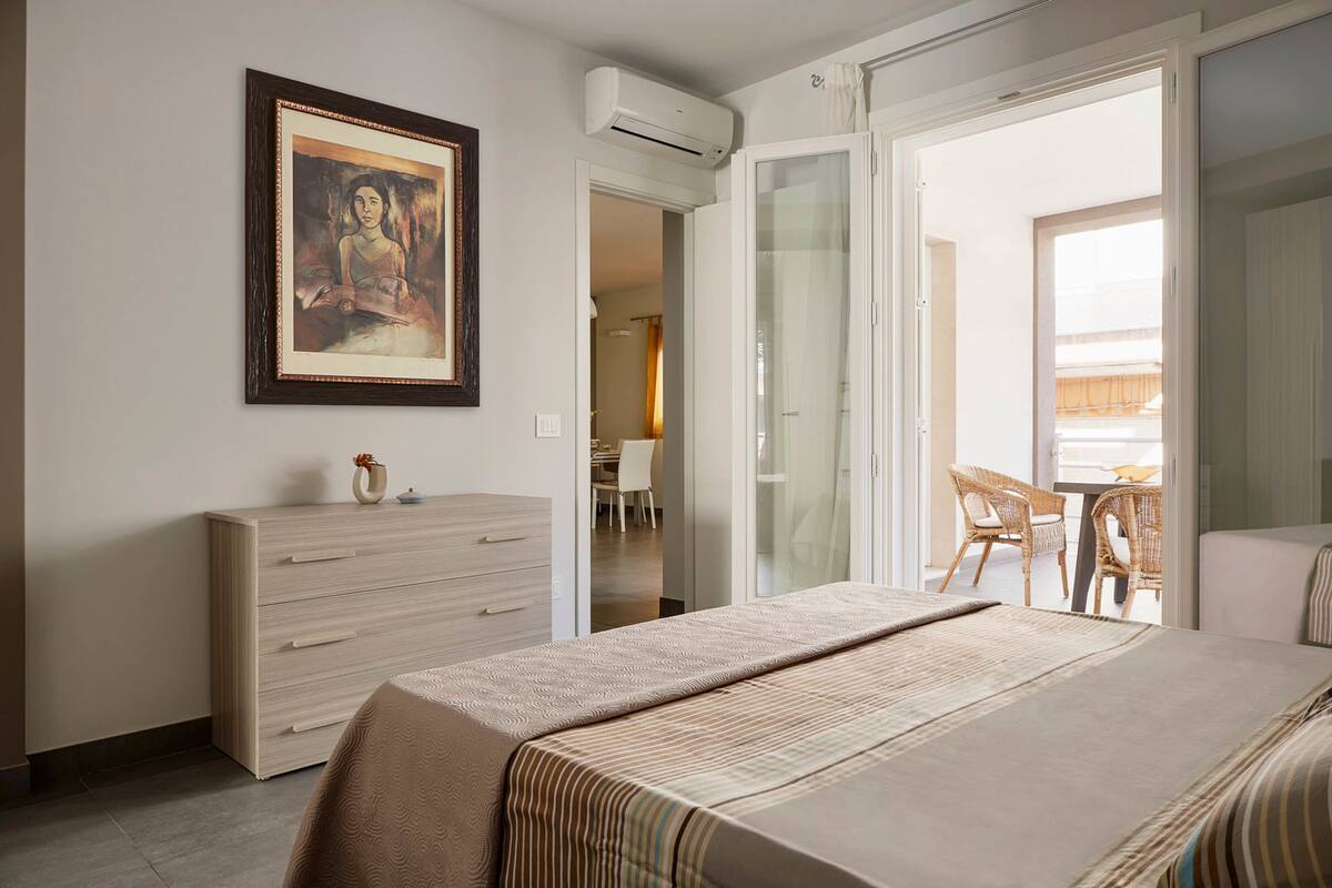 A bedroom with a bed, a dresser, a framed artwork, and a sliding glass door leading to a balcony with wicker furniture.