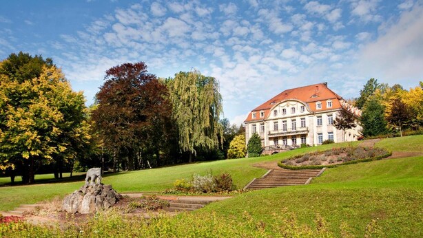 A large, historic building with a red roof, surrounded by a well-maintained garden and a statue. - Hotel Gersfelder Hof (Gersfeld)
