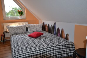 A bedroom with a checkered bedspread, a red pillow, a window with a plant, and a radiator. - pur NATUR & berlin ALEX 40min. (Steinhoefel)