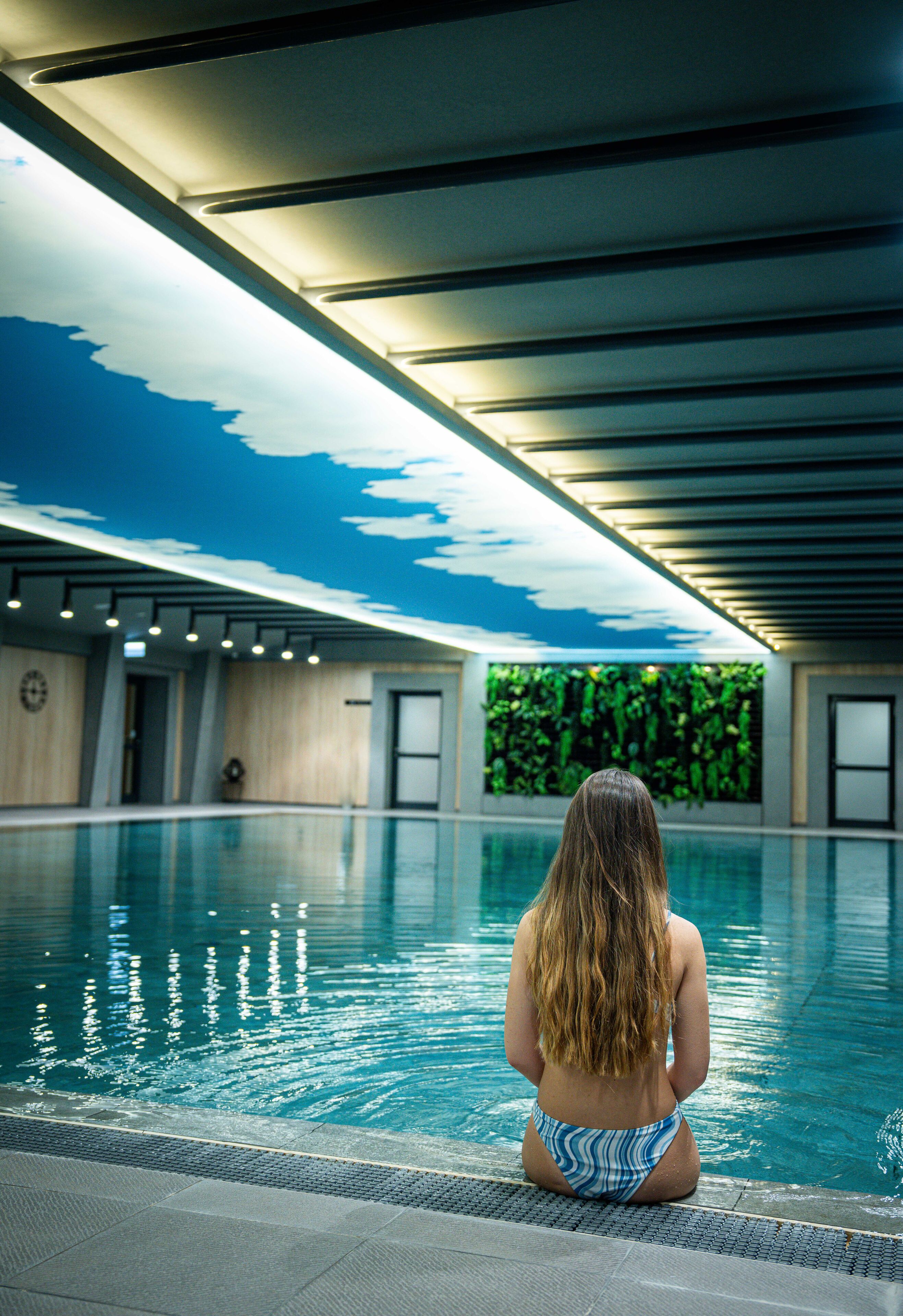 Eine Frau sitzt an einem Innenpool, dessen Decke ein Wolkenfresko und dessen Wand üppiges Grün zeigt.