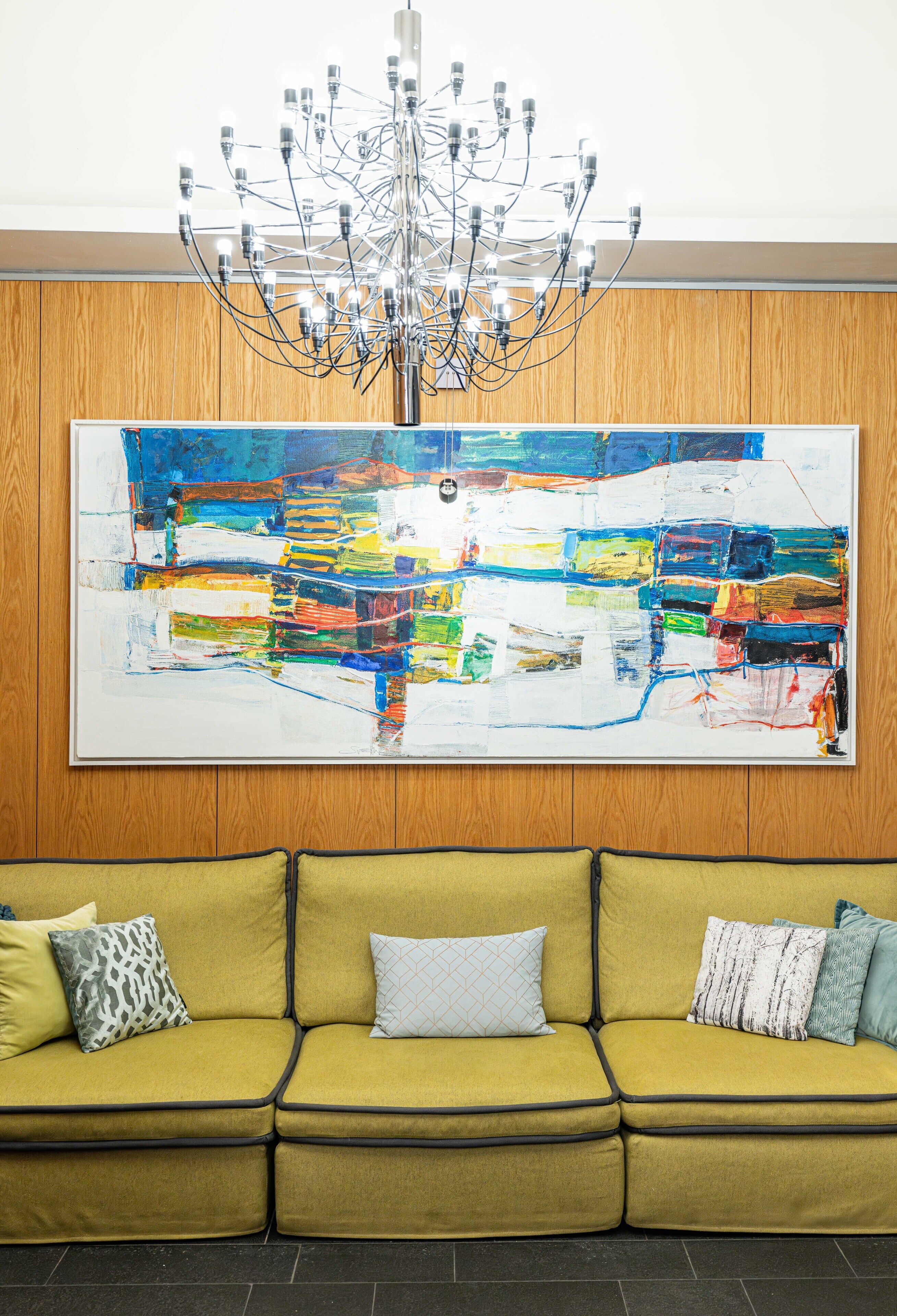 Ein modernes Interieur mit einem großen, abstrakten Gemälde, einer außergewöhnlichen Chandelier und einem senffarbenen Sofa mit gemusterten Kissen.