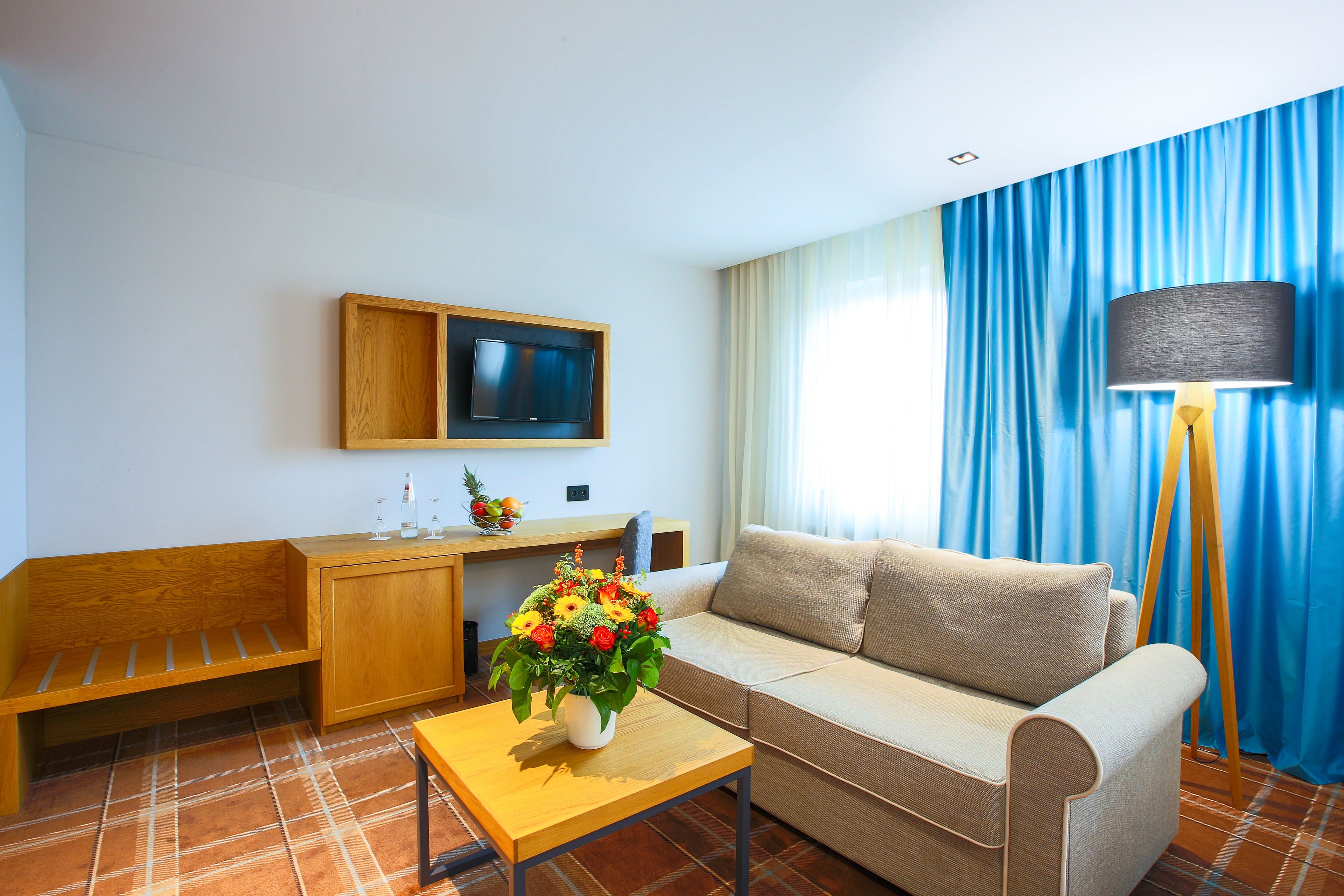 A modern living room with a sofa, a wooden bench, a small table with a vase of flowers, a television on a wooden shelf, and a floor lamp.