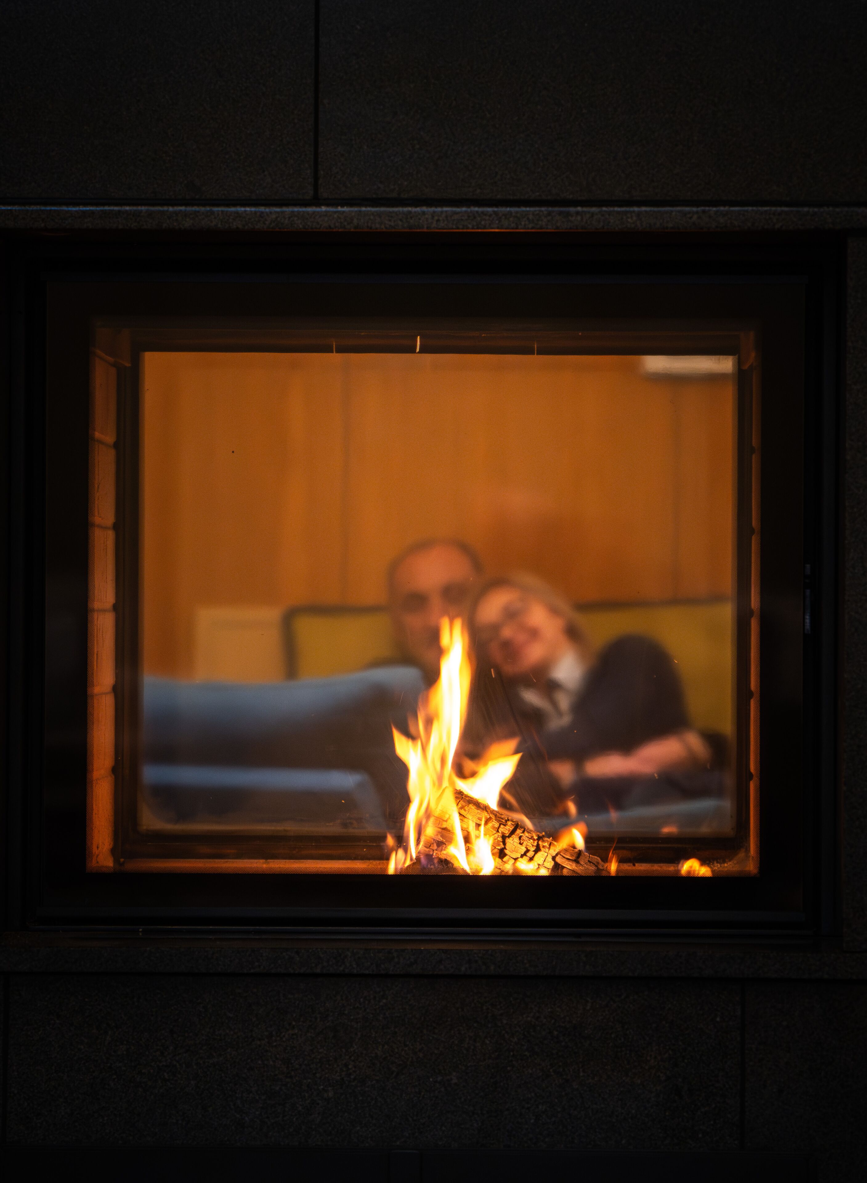 A fireplace with a fire, a couch, and two people sitting in the background.
