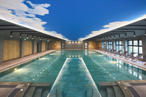 A large indoor swimming pool with a clear view of the sky through the ceiling, surrounded by lounge chairs and a modern architectural design. - Grand Hotel Suhl (Suhl)