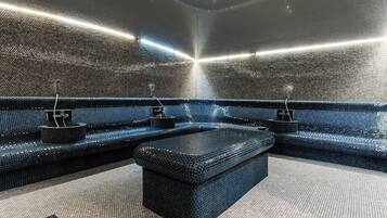 Una habitación moderna, con poca luz, que presenta un original diseño de pared con mosaicos, un área de descanso central y una pequeña mesa con fuente de agua.