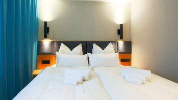 Una cama doble bien hecha con sábanas y toallas blancas, cabecera de madera y dos lámparas de lectura fijadas a la pared.