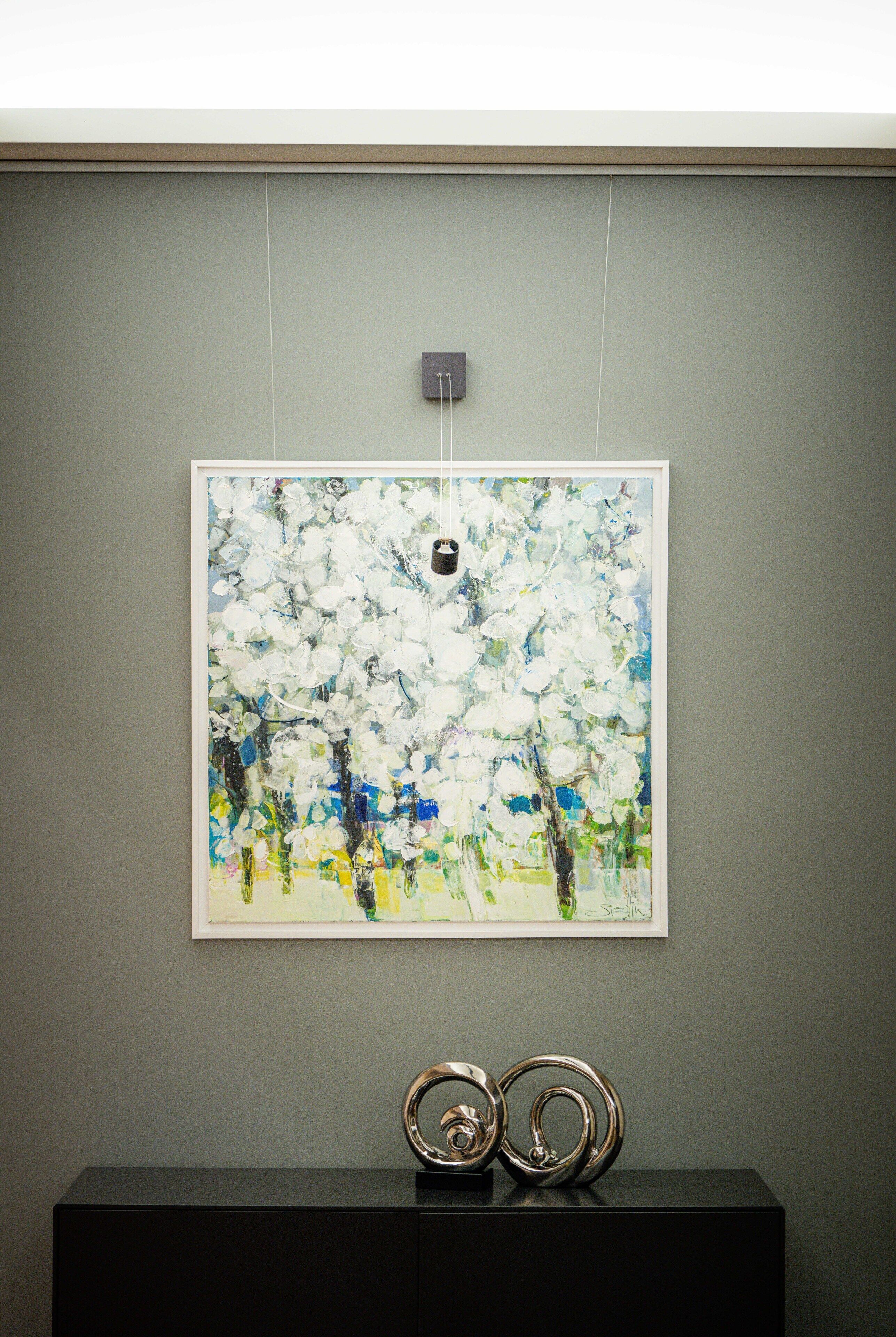 A framed artwork of a tree with white flowers on a wall, a black console table below, and a light switch above.