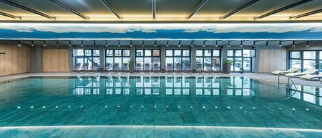 A large indoor swimming pool with clear water, surrounded by lounge chairs and large windows offering views of the outside.