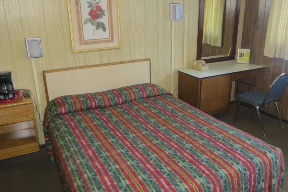 Quarto de hotel com uma cama, uma escrivaninha, uma cadeira, uma cafeteira e um quadro pendurado na parede.