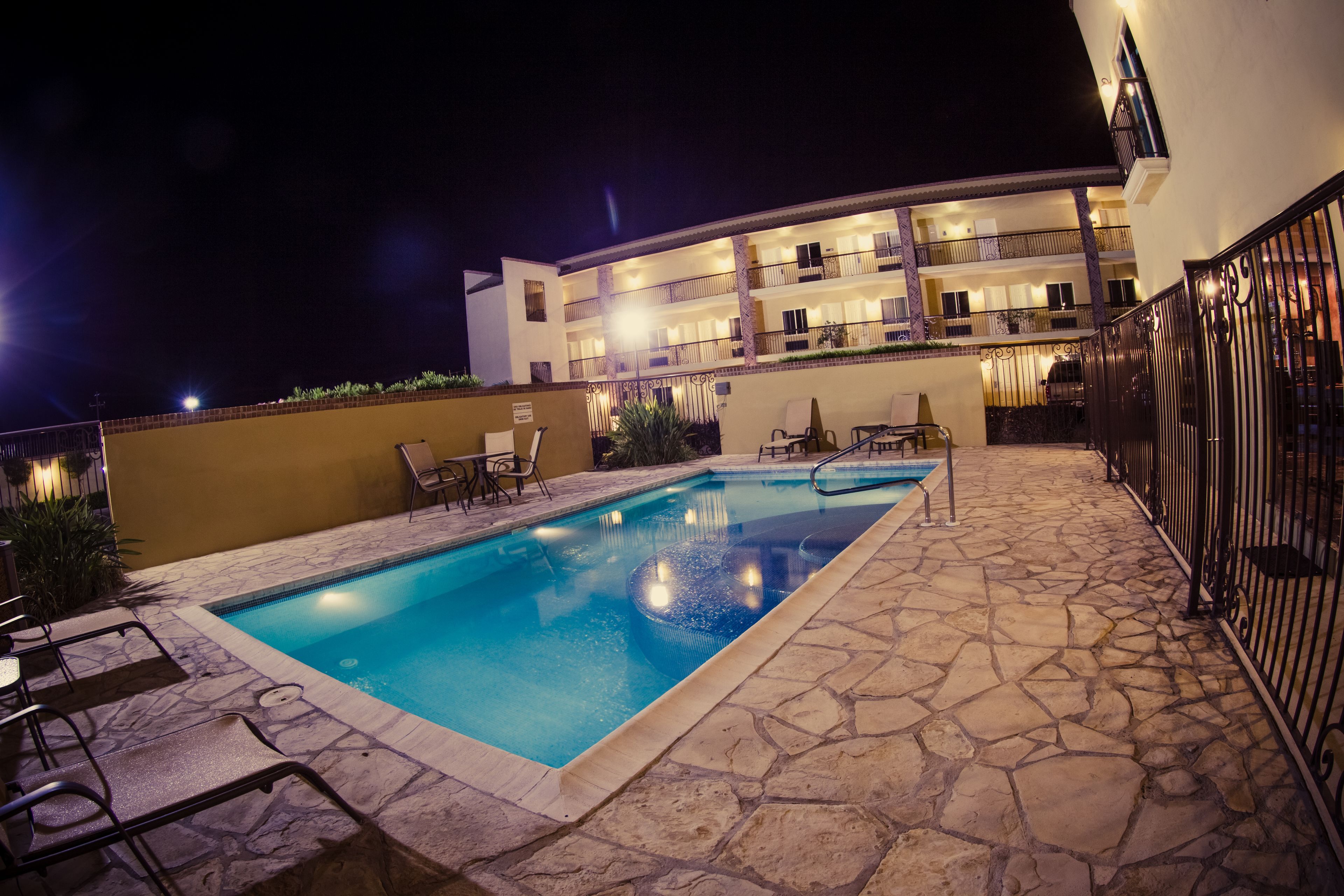 A swimming pool with stone tiles, lounge chairs, and a building with balconies in the background.