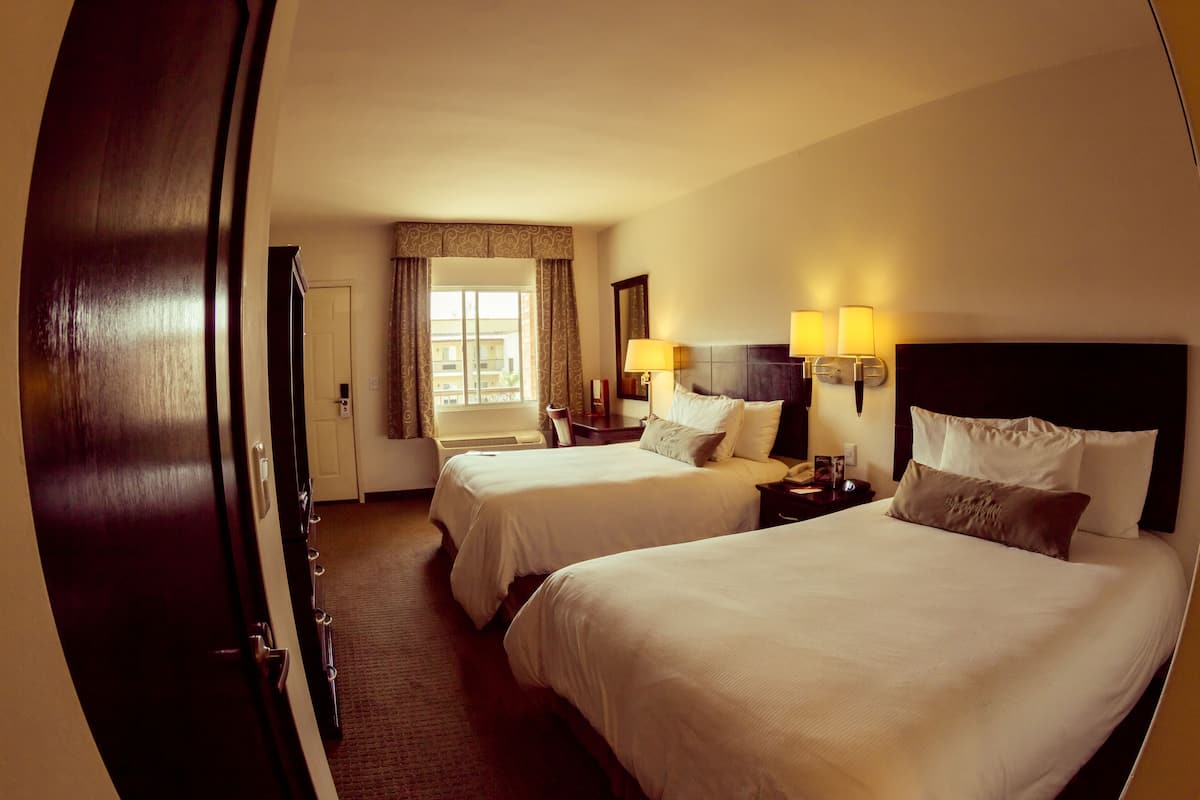 A hotel room with two beds, a window with curtains, a nightstand with lamps, and a door.