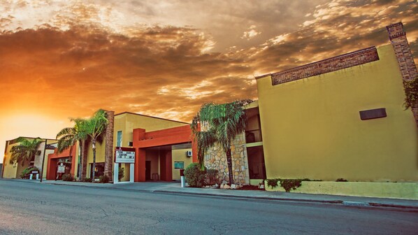 A row of yellow buildings with stone facades and palm trees, a street in the foreground, and a sunset sky.