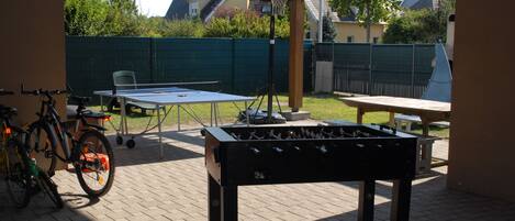 An outdoor recreational area with a foosball table, a ping pong table, and a bicycle.