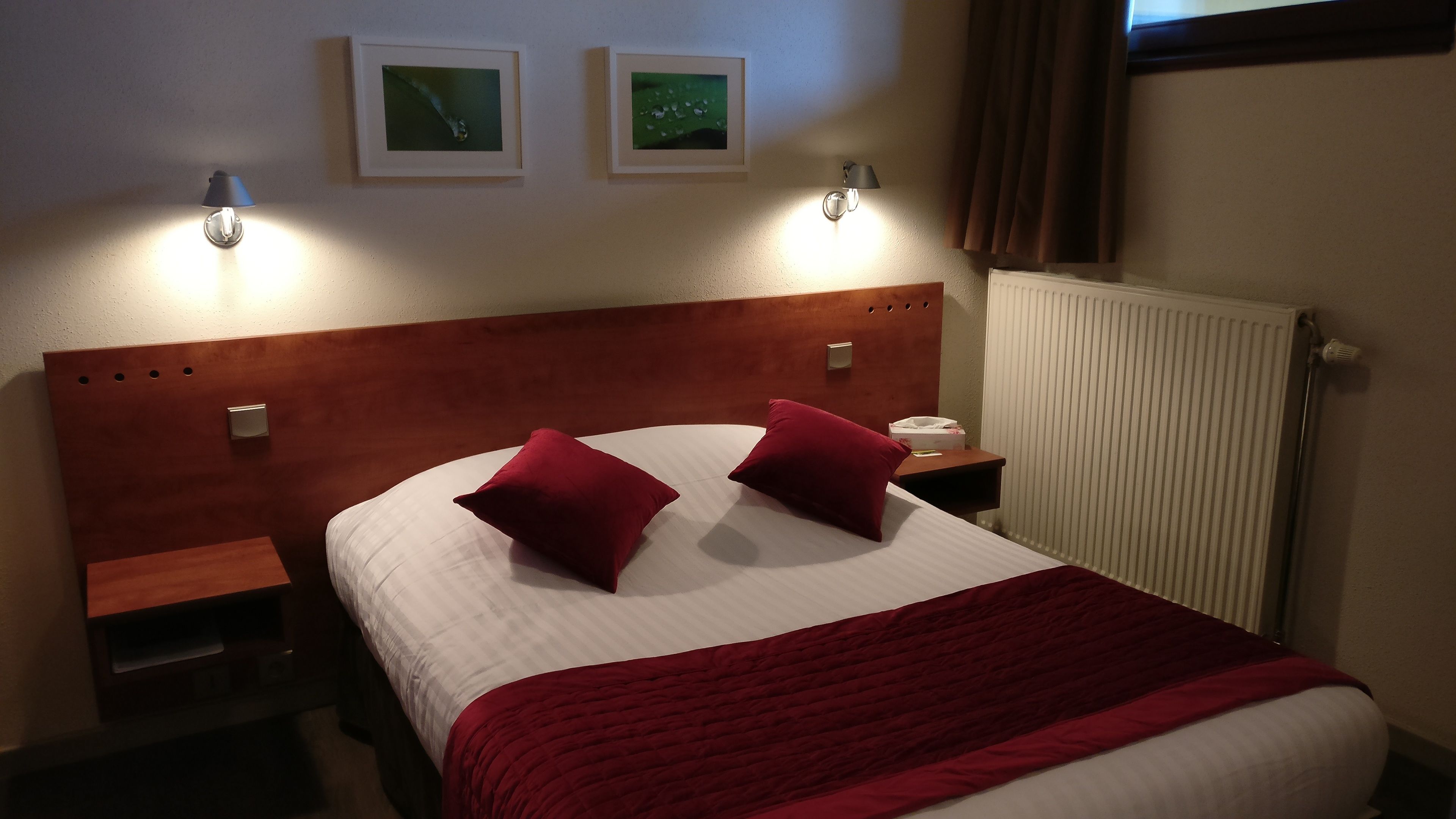 A hotel room with a wooden headboard, two red pillows, a white bedspread, two wall-mounted lamps, and a radiator.