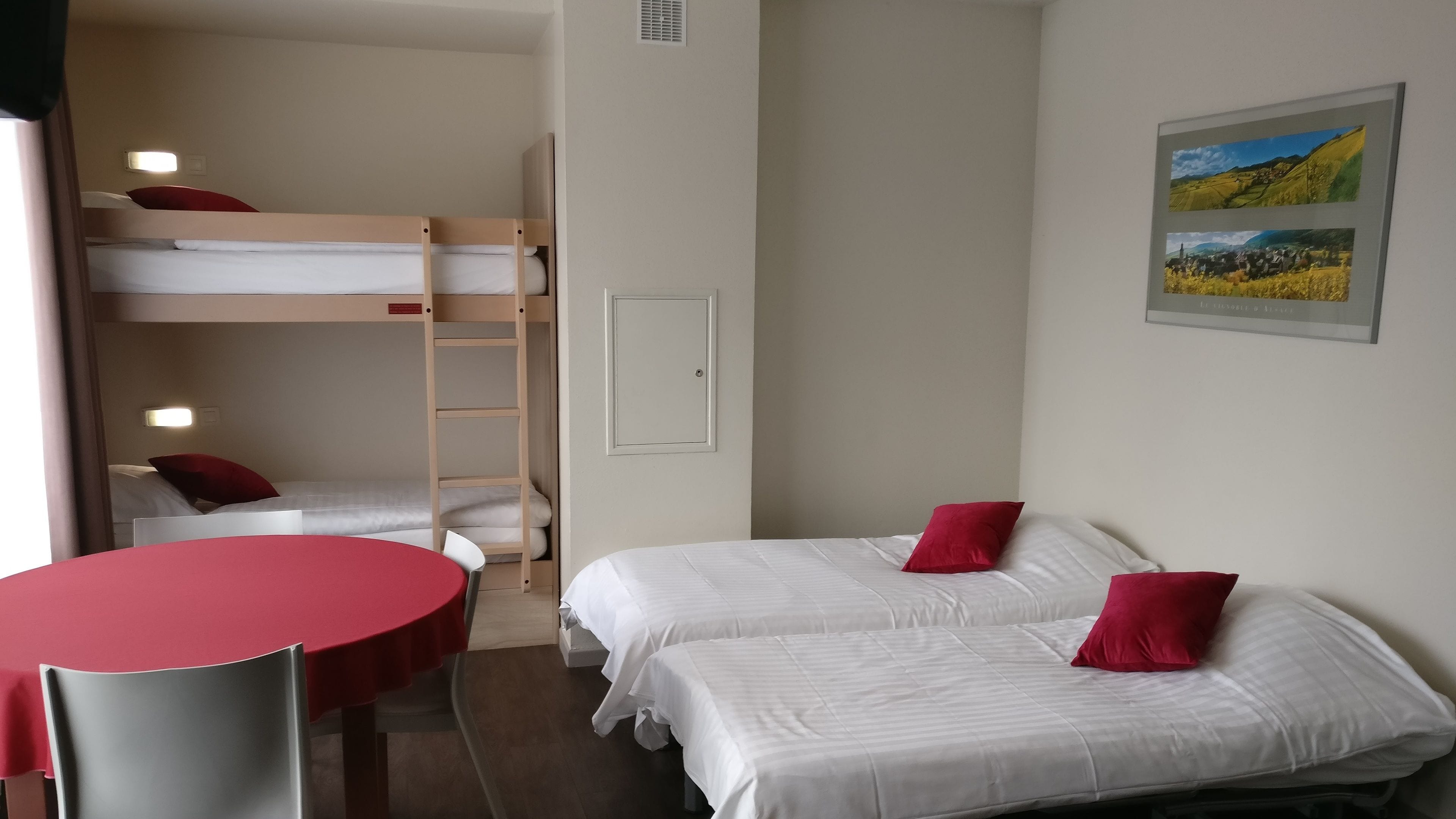 Une chambre d’hôtel compacte comprenant des lits superposés, un lit simple, une petite table et une chaise.