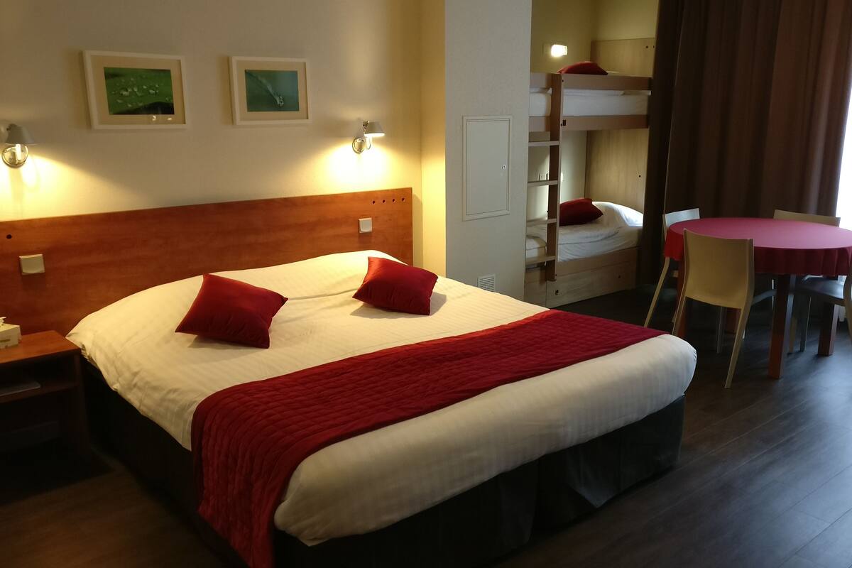 Une chambre d’hôtel avec un grand lit, deux lampes de chevet, une couverture rouge, une petite table avec une chaise, et un lit superposé à l’arrière-plan.