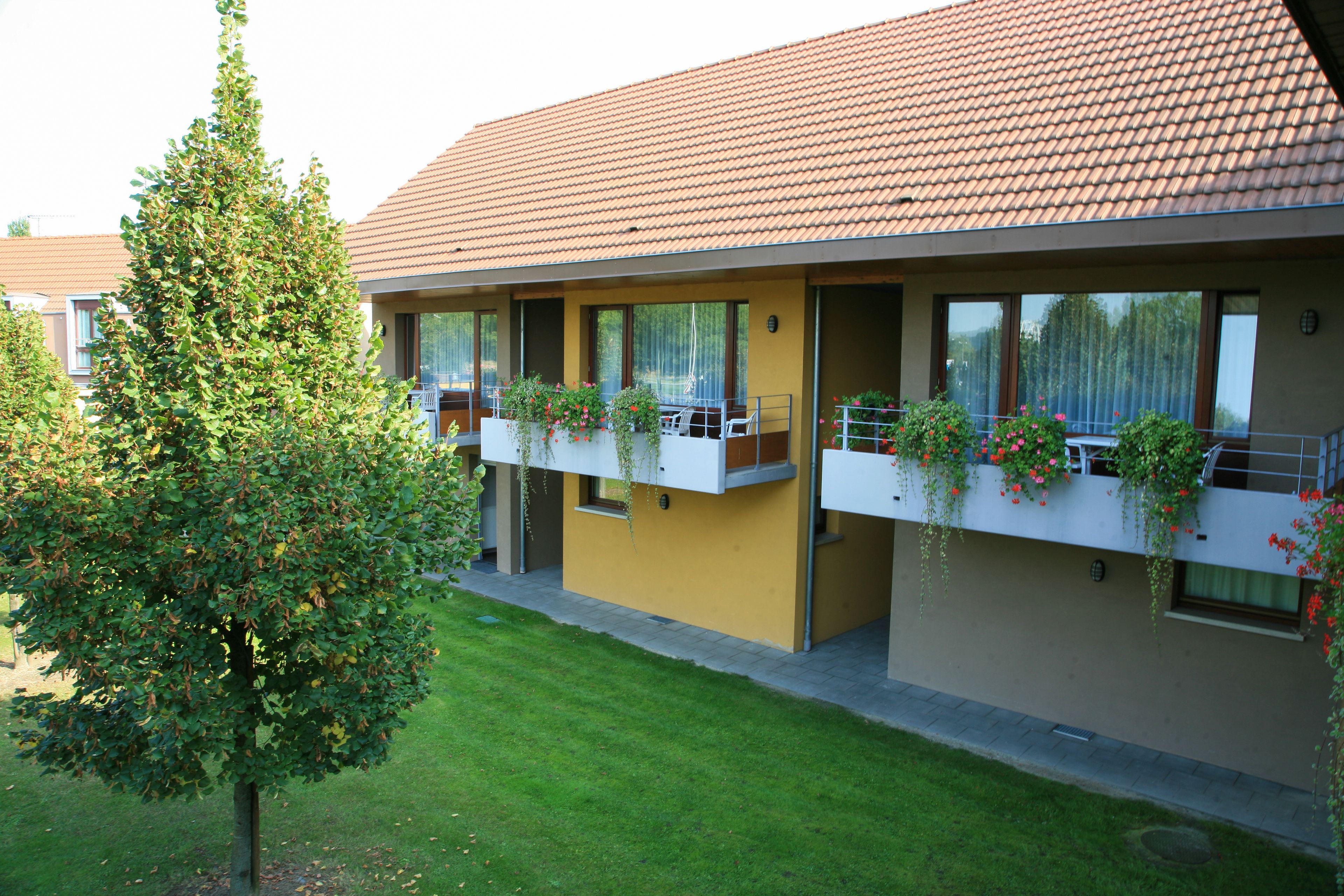 A modern building with a yellow facade and a brown tiled roof. It features balconies with potted plants and a well-maintained lawn.