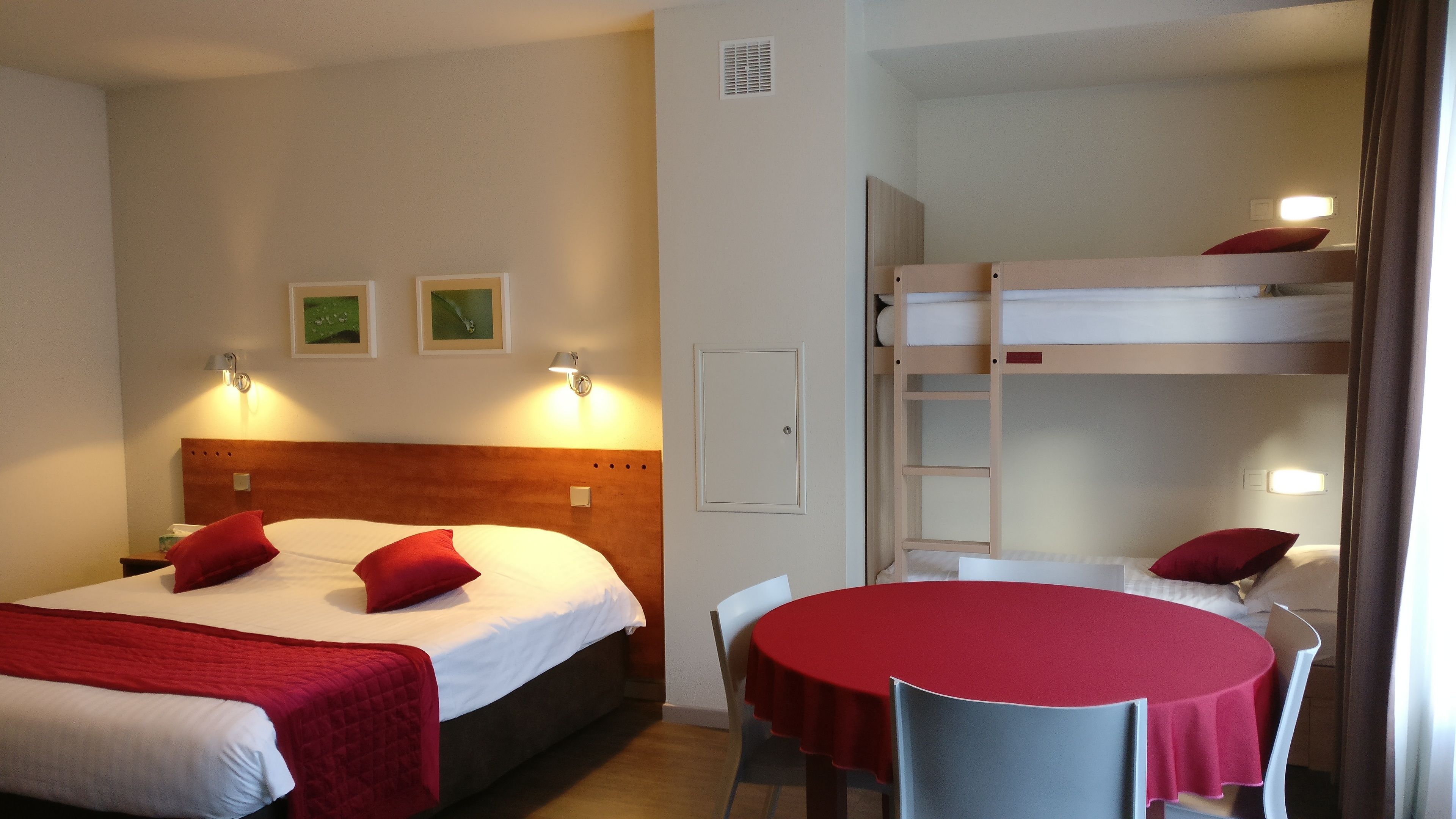 Hotellihuone, jossa on yksi sänky, kerrossänky, pyöreä pöytä tuoleineen ja kaksi seinään kiinnitettyä valaisinta.