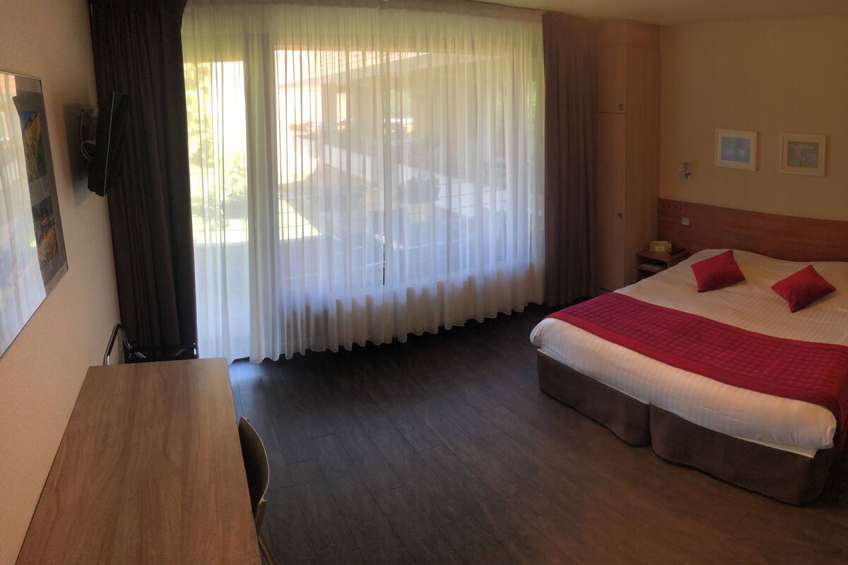 Une chambre d’hôtel avec un grand lit, un bureau en bois, une fenêtre avec des rideaux légers et une vue sur l’extérieur.