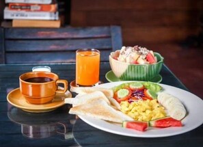 A breakfast setting with a plate of scrambled eggs, a slice of banana, and a salad, a glass of orange juice, and a cup of coffee on a table. - Pai Homey (Pai)