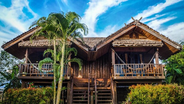 Una casa a due piani in legno con un balcone su ogni piano, circondata da una rigogliosa vegetazione.