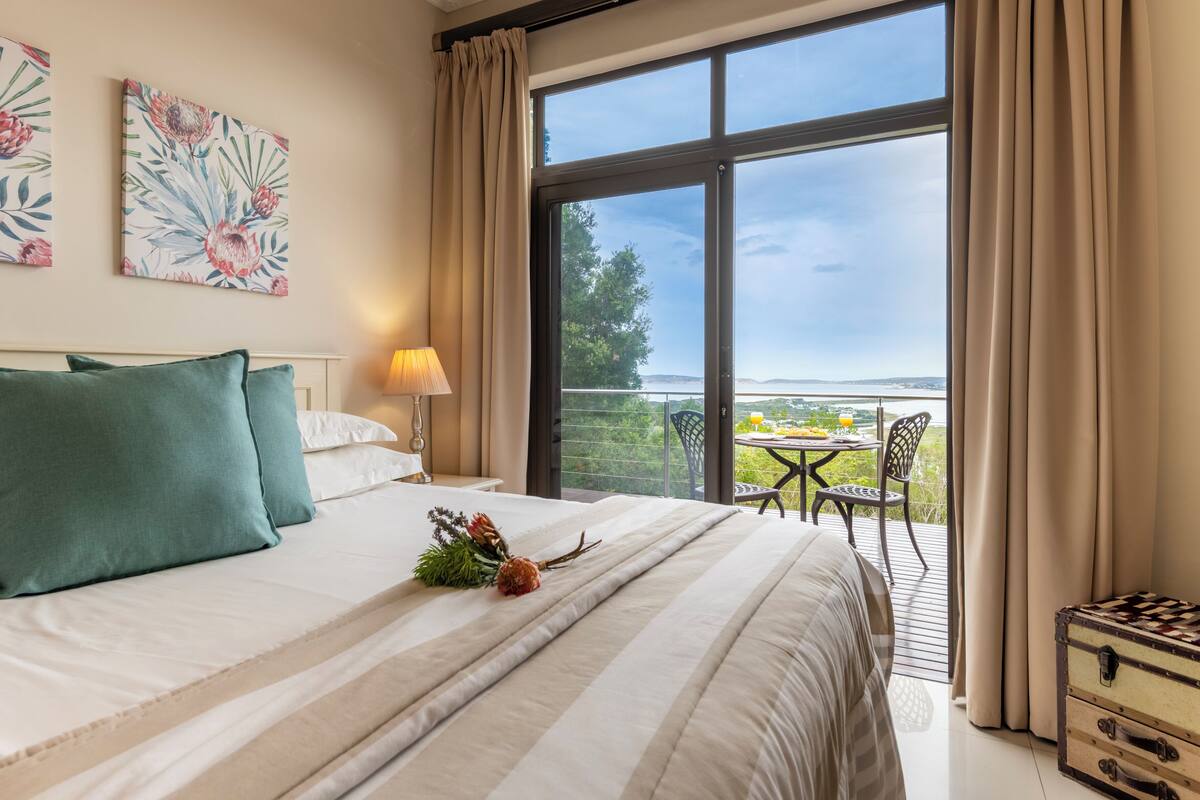 A hotel room with a large bed, a balcony with a table and chairs, and a scenic view.