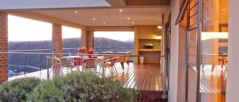 Varanda com mesa e cadeiras, vista para as montanhas e porta de vidro que dá para um quarto.