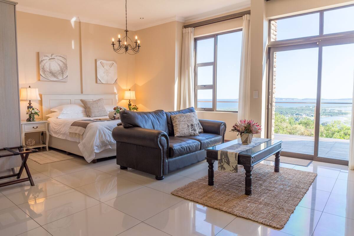 A bedroom with a bed, a dark leather sofa, a wooden coffee table, and a view of the outdoors through large windows.