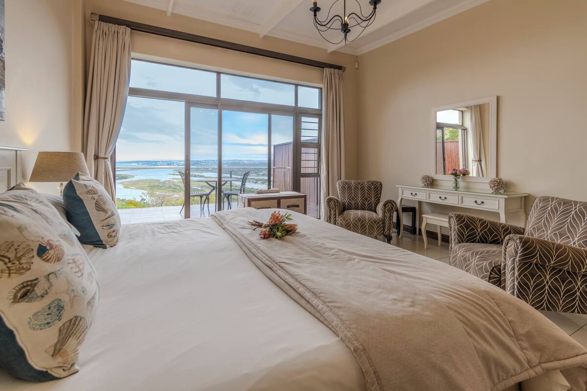 A hotel room with a large bed, a desk, two chairs, a mirror, and a view of the outdoors.