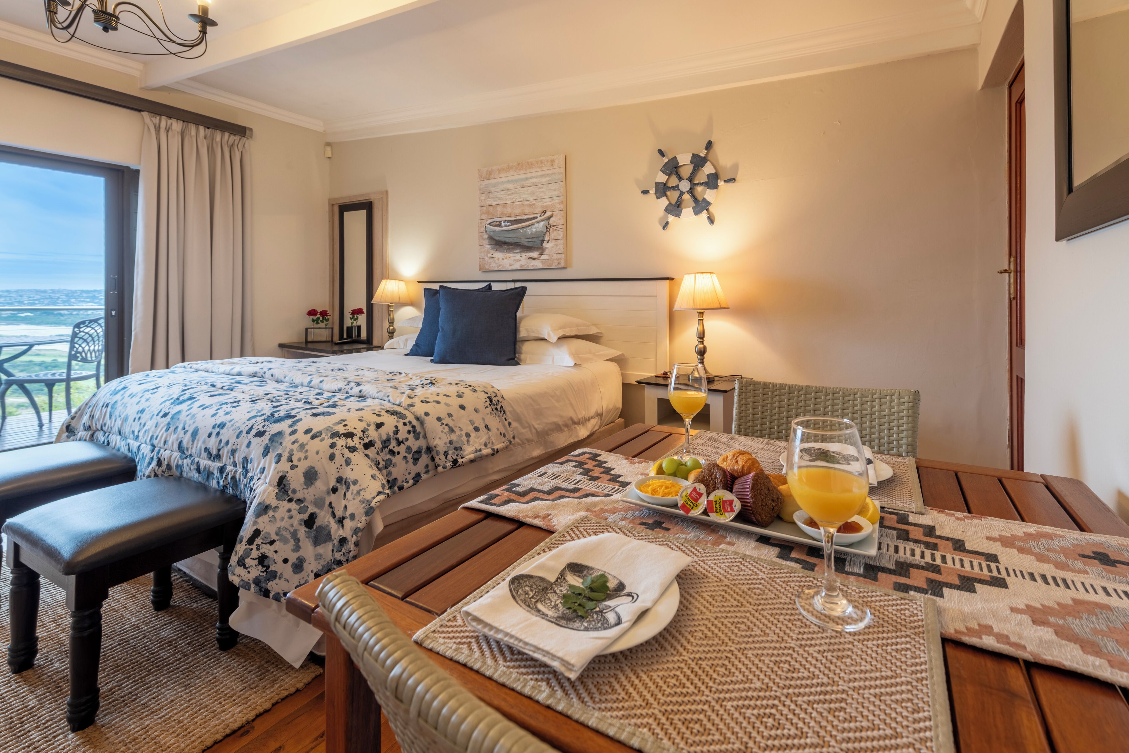 A hotel room with a bed, a dining table with breakfast, a view of the ocean, and a ship decoration on the wall.